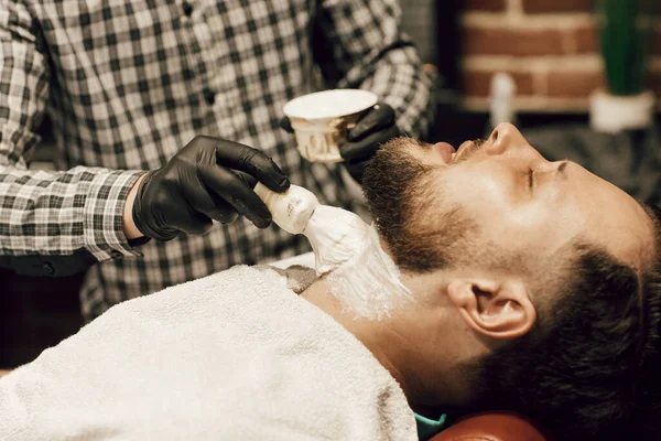 Jovem barbudo sentado na cadeira da barbearia. Cabeleireiro lubrifica a barba com gel de barbear — Fotografia de Stock