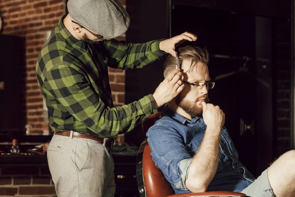 O cabeleireiro ajusta o corte de cabelo de clientes com a tesoura. Um jovem com barba sentado em uma cadeira em uma barbearia — Fotografia de Stock