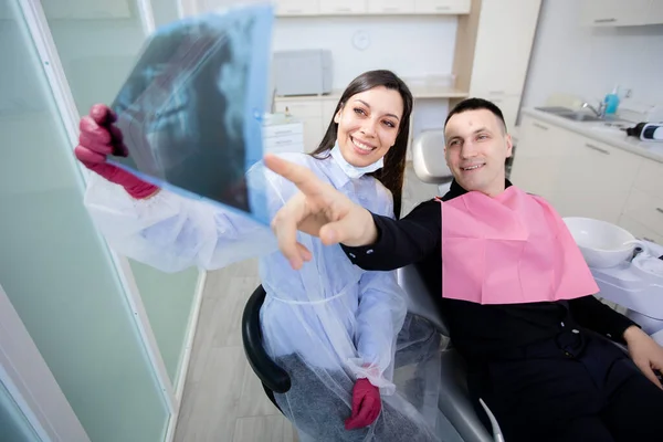 Sorridente dentista femminile e giovane uomo seduto in ufficio a guardare i raggi X. Il dentista mostra al paziente una radiografia — Foto Stock