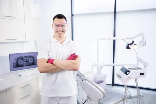 Portrait of a young smiling dentist in work glasses and in a white uniform. The man folded his arms and stands at the dental chair in the office — Stock Photo, Image