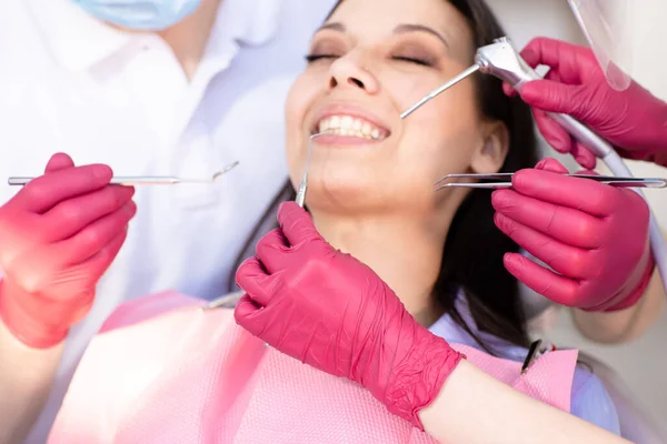 Primo piano mani di due dentisti in guanti rosa con strumenti e sorridente giovane donna su una sedia dentale sullo sfondo. Sorriso sano e bianco, concetto di odontoiatria — Foto Stock