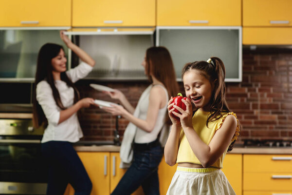 Happy girl with eyes closed smiling and holding sweet pepper. Girl standing in the kitchen and playing with a big red paprika. In the background, two women stack dishes. Advertising healthy eating Stock Image