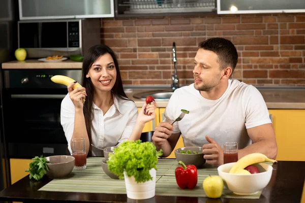 Pasangan muda yang bahagia tersenyum memasak bersama di rumah. Pria dan wanita duduk di meja, berbicara, bersenang-senang, makan sayuran. Konsep makan yang sehat, hubungan — Stok Foto