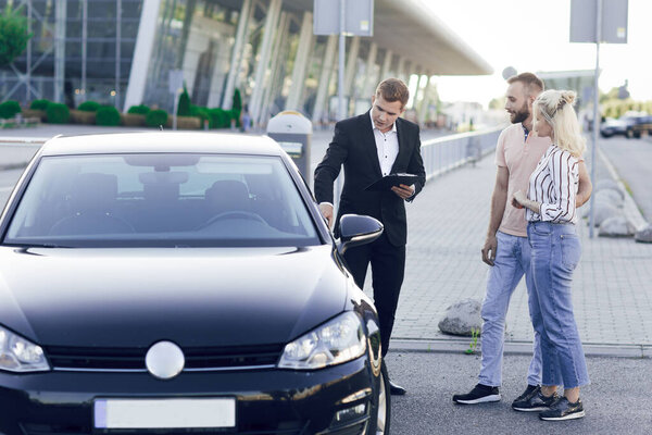 A young car dealer in a business suit shows buyers a new car. Young couple, man and woman, buy a car. Purchase of machines, test drive Royalty Free Stock Photos