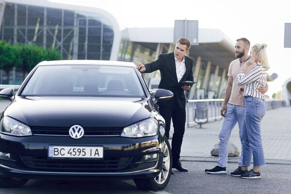 A young car dealer in a business suit shows buyers a new car. Young couple, man and woman, buy a car. Purchase of machines, test drive Stock Picture