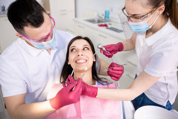 Dental treatment during coronavirus quarantine. Smiling young woman in a dental chair. Dentist and assistant in protective masks and gloves check the patients teeth Stock Photo