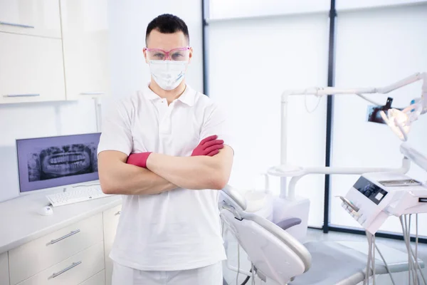 Korunaklı maske takan, gözlüklü ve beyaz üniformalı genç bir dişçinin portresi Coronavirus karantinası sırasında ofiste duruyor. Acil servis. — Stok fotoğraf