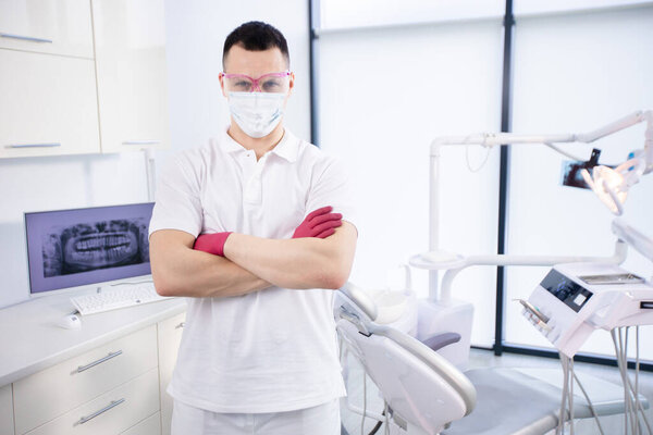 Portrait of a young dentist in protective mask, work glasses and in a white uniform stands in the office during coronavirus quarantine. Emergency care Royalty Free Stock Images