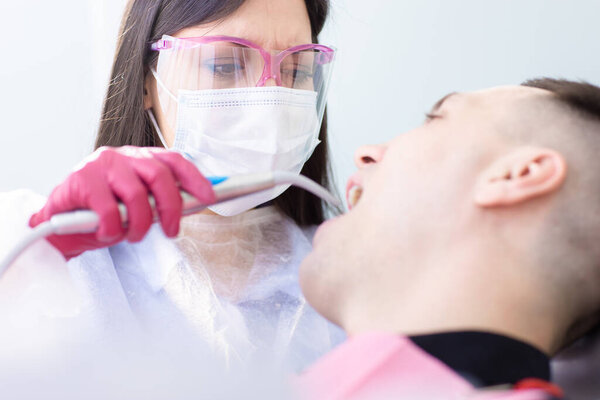Close up view of female dentist in protective mask and glasses examines a male patient. Young man in the dentists chair. Visit to the dentist during a coronavirus quarantine Stock Photo