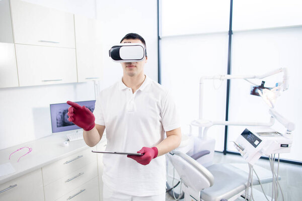 A male dentist in uniform stands in the office and checks the digital image before the patients visit. The latest technologies in dentistry, 3D images, virtual reality Stock Photo