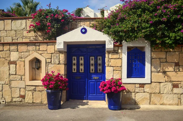 Eski taş bina (ev) mavi ahşap kapılı ve pencereli. Önünde güzel pembe çiçekler var. Geleneksel Bodrum Ege mimari biçimi 