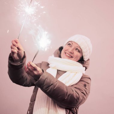 Kış kıyafetleri içinde yetişkin bir kadın pembe bir duvarın arkasında ışıldayan kıvılcımlar tutuyor.