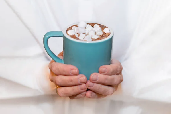An adult woman in a white shirt holds a blue cup of cocoa with marshmallows in her hands. — Stock Photo, Image