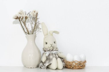 Paskalya kompozisyonu, yumuşak tavşan iguşkası sepette beyaz yumurta ve odadaki rafta bir vazo çiçek.