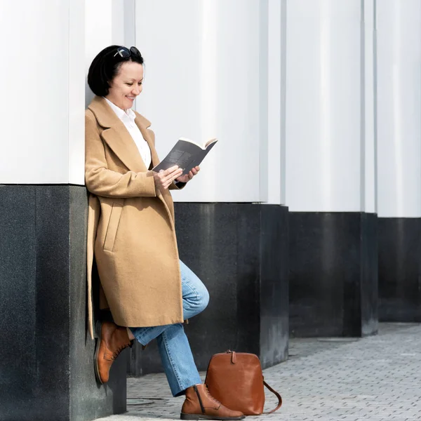 Dorosła kobieta modnie ubrana stoi na ulicy w pobliżu budynku czytającego książkę.. Obrazy Stockowe bez tantiem