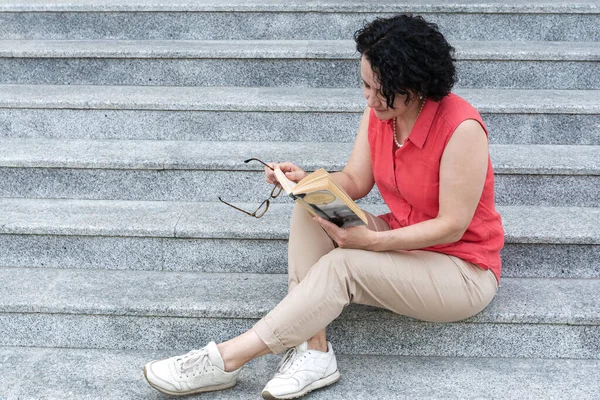 Una mujer se sienta en las escaleras de la ciudad y lee un libro. — Foto de Stock