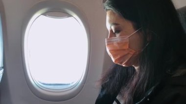 Koruyucu maskeli koyu renk saçlı bir kadın pencerenin yanındaki kulübede oturup kitap okuyor..