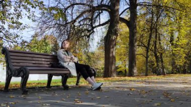Genç bir kadın sonbahar parkında bir bankta oturuyor ve güzel sarı yapraklara bakıyor..