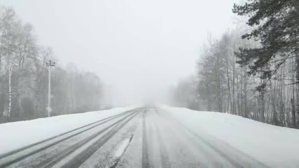 Vue à travers le pare-brise d'une voiture sur une route enneigée et glacée vide en hiver sous de fortes chutes de neige avec une mauvaise visibilité dans la forêt. — Video