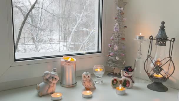 Una vista de la composición navideña de velas de muñecos de nieve de abeto y búhos en el alféizar de la ventana de la casa con una vista de los árboles cubiertos de nieve fuera de la ventana. — Vídeo de stock