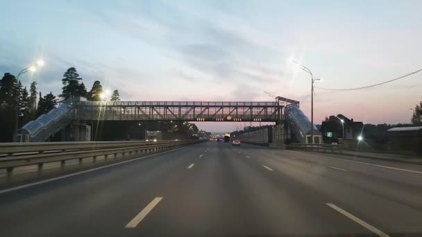 Vue de l'autoroute et du passage supérieur pour piétons à travers le pare-brise d'une voiture le soir sous la lumière des lanternes. Mouvement lent. — Video