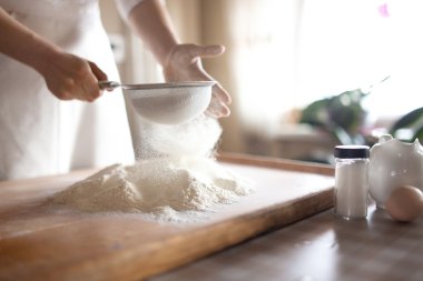 young woman sifting flour into bowl at the kitchen clipart