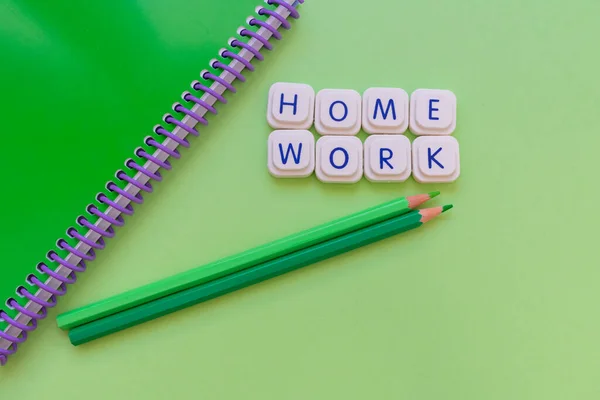 Home work message made with board game letters, with a green notebook and two green pencils, over a green background. Studies and work concept