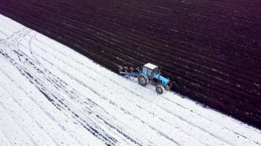 Mavi bir traktör karla kaplı bir tarlayı sürüyor. Traktörün arkasında kara toprak var. Rusya, Ural, Hava Görünümü  