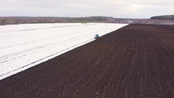 Dolly ráközelít. Egy kék traktor felszántja a havas mezőt. A traktor mögött fekete föld van. Oroszország, Ural — Stock videók