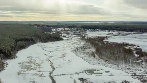 Sorvolando una palude ricoperta di neve con una striscia scura del fiume. Il villaggio sullo sfondo. Le luci di un sole. 4K — Video Stock