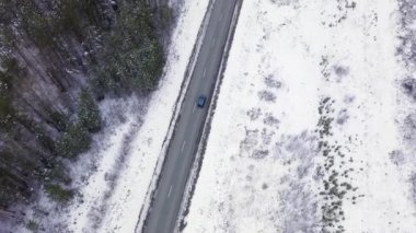 Mavi bir araba kış asfalt yolu boyunca gidiyor. Yol elektrik hatları boyunca uzanıyor. Ağaçlarda ve yol kenarlarında kar. 4K