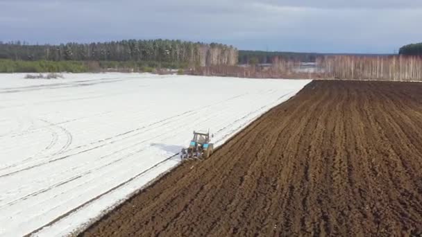 Долли Зум. Синий трактор вспахивает поле, покрытое снегом. За трактором чернозем. Russia, Novosibirsk — стоковое видео
