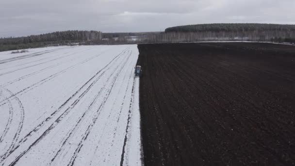 A blue tractor plows a field covered with snow. Behind the tractor is black earth. Russia, Ural. 4K — Stock Video