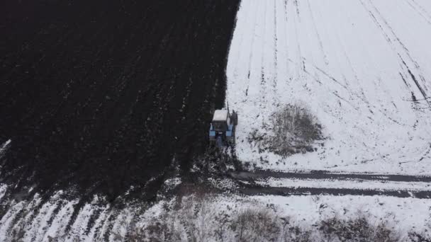 Un tractor azul arada un campo cubierto de nieve. Detrás del tractor hay tierra negra. El borde del campo, comience. Rusia, Ural. 4K — Vídeo de stock