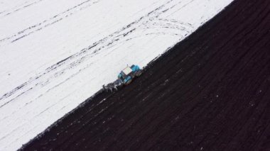 Mavi bir traktör karla kaplı bir tarlayı sürüyor. Traktörün arkasında kara toprak var. Alanın kenarına. Rusya, Ural. 4K