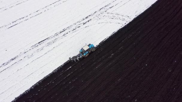Синий трактор вспахивает поле, покрытое снегом. За трактором чернозем. Край поля. Россия, Урал. 4K — стоковое видео