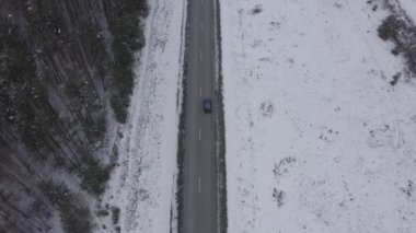 Mavi bir araba kış asfalt yolu boyunca gidiyor. Yol elektrik hatları boyunca uzanıyor. Ağaçlarda ve yol kenarlarında kar. 4K