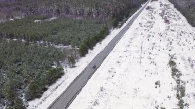 Mavi bir araba kış asfalt yolu boyunca gidiyor. Yol elektrik hatları boyunca uzanıyor. Ağaçlarda ve yol kenarlarında kar. 4K