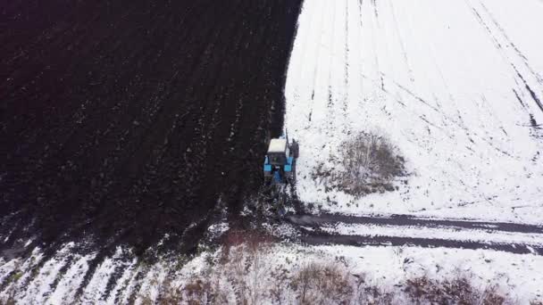 Een blauwe trekker ploegt een veld bedekt met sneeuw. Achter de trekker is zwarte aarde. Begin aan de rand van het veld. Rusland, Oeral. 4K — Stockvideo