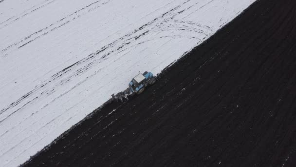 Modrý traktor orá pole pokryté sněhem. Za traktorem je černá země. Okraj pole. Rusko, Ural. 4K — Stock video