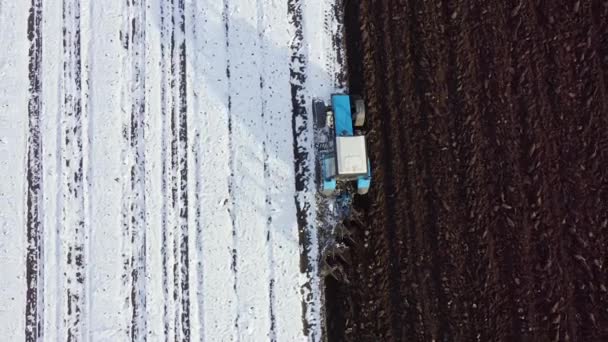Um trator azul limpa um campo coberto de neve. Atrás do trator está a terra negra. Rússia, Ural. 4K — Vídeo de Stock