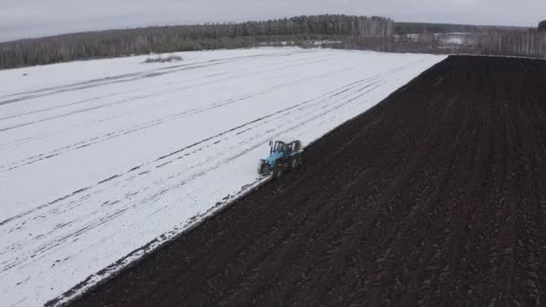 Een blauwe trekker ploegt een veld bedekt met sneeuw. Achter de trekker is zwarte aarde. Rusland, Oeral. 4K — Stockvideo