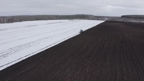 Een blauwe trekker ploegt een veld bedekt met sneeuw. Achter de trekker is zwarte aarde. Rusland, Oeral. 4K — Stockvideo