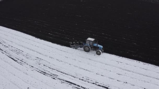 Modrý traktor orá pole pokryté sněhem. Za traktorem je černá země. Rusko, Ural. 4K — Stock video