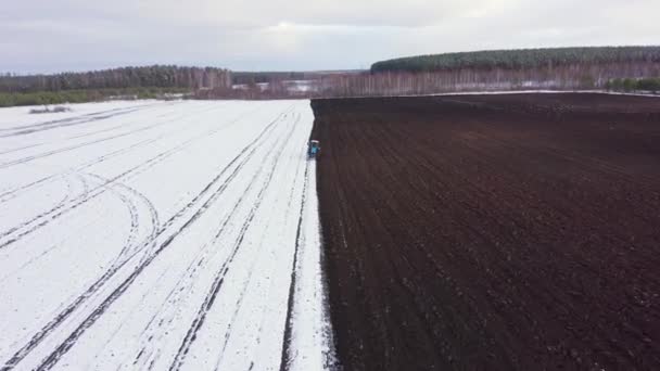 Dolly ráközelít. Egy kék traktor felszántja a havas mezőt. A traktor mögött fekete föld van. Oroszország, Ural — Stock videók