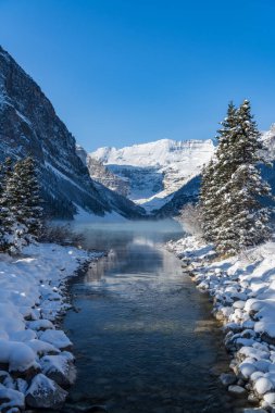 Louise Gölü kışın erken saatlerinde güneşli bir günde. Turkuaz renkli su yüzeyinde süzülen sis. Açık mavi gökyüzü, arka planda karla kaplı dağlar. Banff Ulusal Parkı 'nda güzel doğal manzara.
