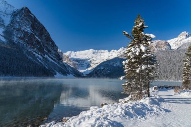 Louise Gölü kışın erken saatlerinde güneşli bir günde. Turkuaz renkli su yüzeyinde süzülen sis. Açık mavi gökyüzü, arka planda karla kaplı dağlar. Banff Ulusal Parkı 'nda güzel doğal manzara.