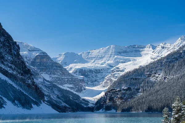 Lake Louise an einem sonnigen Frühwinter-Morgen. Nebel schwebt auf türkisfarbener Wasseroberfläche. Klarer blauer Himmel, im Hintergrund schneebedeckte Berge. Schöne Naturlandschaft im Banff Nationalpark. — Stockfoto