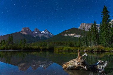 Üç Kız Kardeş üçlüsünün tepesindeki yıldızlarla dolu gece, yıldızlı gökyüzü derenin su yüzeyine yansıyor. Banff Ulusal Parkı, Kanada Kayalıkları, Alberta, Kanada.