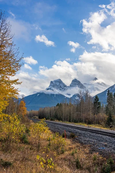 Railway scenery in autumn season, blue sky with white clouds and snow capped The Three Sisters trio of peaks in the background. Canmore, Alberta, Canada.